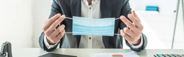 Vista ritagliata di uomo d'affari con maschera medica, seduto sul posto di lavoro su sfondo sfocato, banner — Foto stock