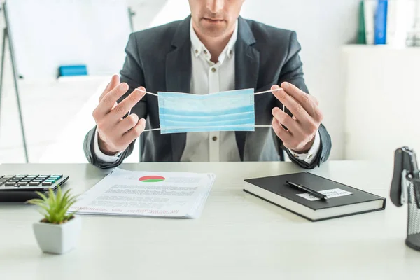 Cropped view of businessman holding medical mask, while sitting at desk on blurred background — Stock Photo