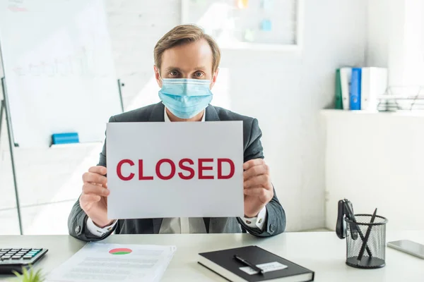 Businessman in medical mask holding sign with closed lettering, while sitting at workplace on blurred background — Stock Photo