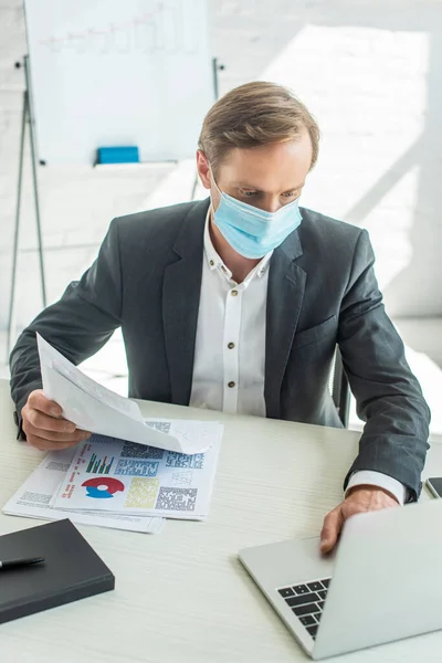 Empresario en máscara médica, sosteniendo papeles y escribiendo en el portátil, mientras está sentado en el lugar de trabajo en un fondo borroso - foto de stock