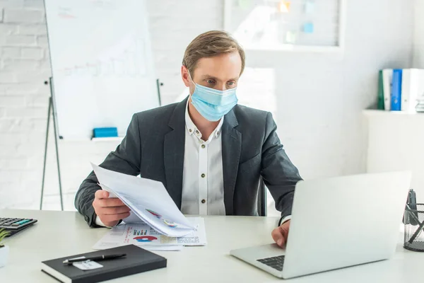 Empresario en máscara médica, sosteniendo papeles y usando computadora portátil, mientras está sentado en el lugar de trabajo en un fondo borroso - foto de stock