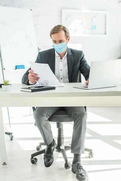 Longitud completa de hombre de negocios en máscara médica mirando el papel, mientras está sentado en la mesa en la oficina sobre fondo borroso - foto de stock