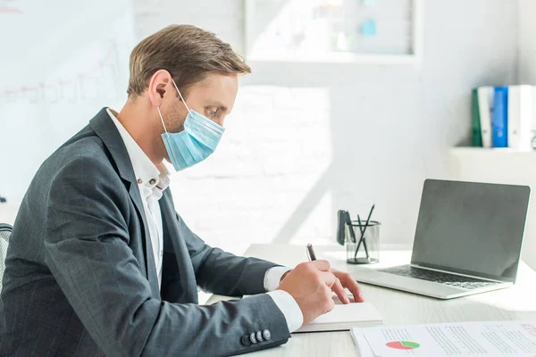 Vue latérale de l'homme d'affaires en masque médical, écrit dans un carnet vierge, tout en étant assis sur le lieu de travail sur fond flou — Photo de stock