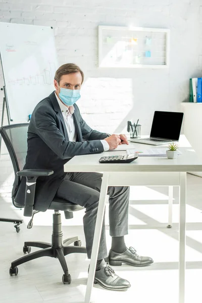 Longitud completa de hombre de negocios en máscara médica mirando a la cámara, mientras que la escritura en el cuaderno, sentado en el lugar de trabajo en la oficina - foto de stock