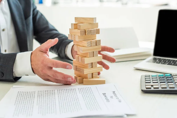 Vista recortada de la quiebra con las manos cerca de bloques juego de torre de madera, sentado en el escritorio sobre fondo borroso - foto de stock