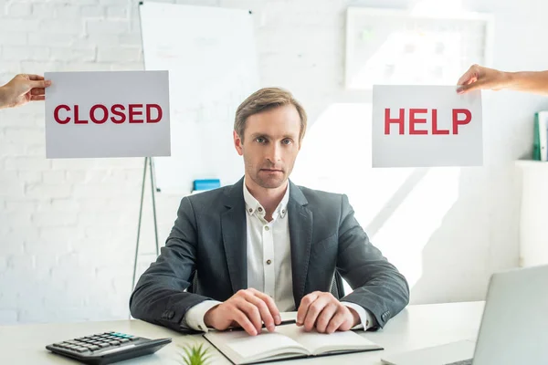 Businessman looking at camera, while sitting at workplace near signs with closed and help lettering on blurred background — Stock Photo
