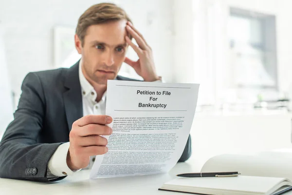 Empresário pensativo olhando para petição de falência, enquanto sentado no local de trabalho em fundo turvo — Fotografia de Stock