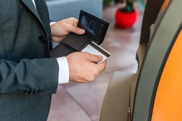 Vista recortada del hombre de negocios que pone la tarjeta de crédito en la cartera, mientras que de pie cerca del cajero automático con la calle borrosa en el fondo - foto de stock