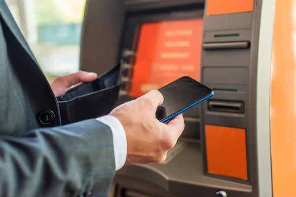 Cropped view of businessman with empty wallet and smartphone standing near atm on blurred background — Stock Photo