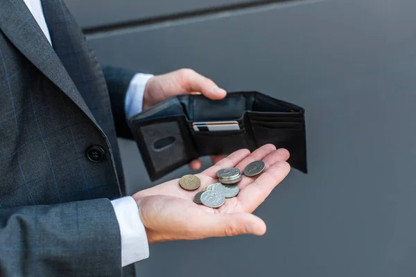 Vista recortada del hombre de negocios con monedas en la palma, sosteniendo la cartera vacía con tarjetas de crédito sobre fondo texturizado gris - foto de stock