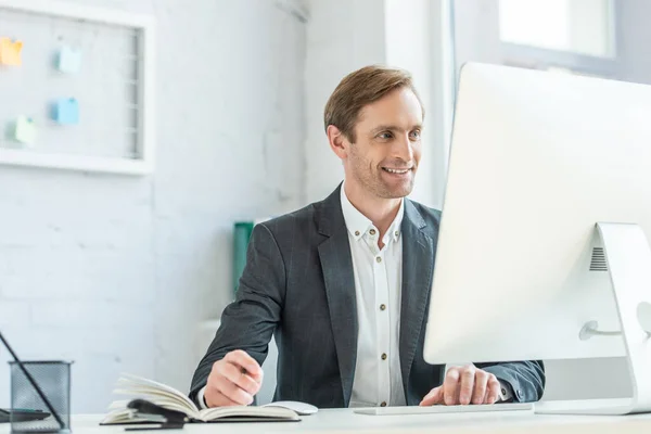 Felice uomo d'affari guardando monitor del computer, mentre seduto sul posto di lavoro in ufficio su sfondo sfocato — Foto stock