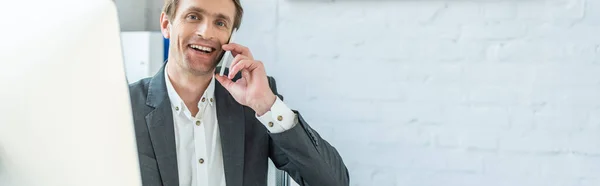 Улыбающийся бизнесмен смотрит в камеру, разговаривая по телефону в офисе с размытым монитором компьютера на переднем плане, баннер — стоковое фото