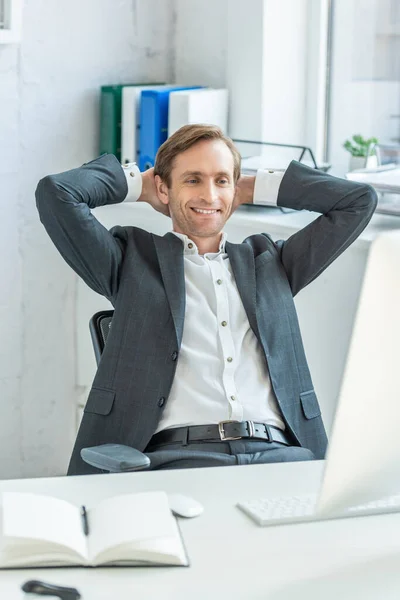 Homem de negócios feliz com as mãos atrás da cabeça, olhando para o monitor do computador enquanto sentado no local de trabalho em primeiro plano turvo — Fotografia de Stock