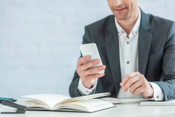 Vista recortada del hombre de negocios positivo usando un teléfono inteligente, mientras está sentado en la mesa sobre un fondo borroso — Stock Photo