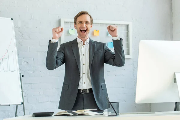 Vista frontal del empresario emocionado con un gesto sí mirando a la cámara, mientras está de pie cerca del lugar de trabajo en la oficina - foto de stock