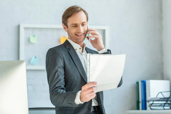 Улыбающийся бизнесмен смотрит на документ в пластиковом файле, разговаривая по телефону с размытым офисом на заднем плане — стоковое фото