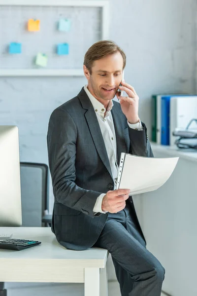 Positiver Geschäftsmann beim Betrachten eines Dokuments, während er auf dem Tisch neben dem Computermonitor auf verschwommenem Hintergrund sitzt — Stockfoto