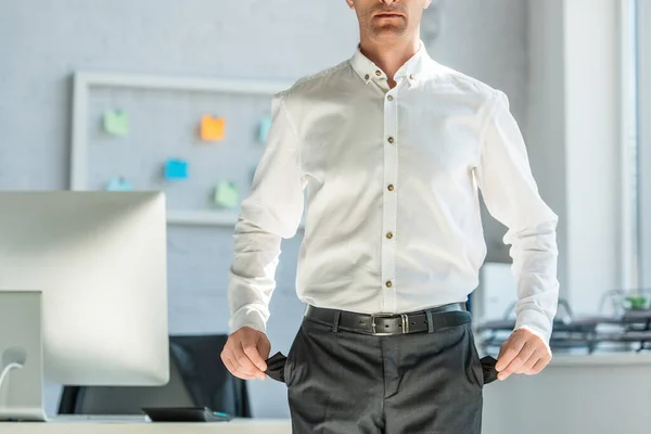 Ausgeschnittene Ansicht eines Geschäftsmannes, der leere Taschen zeigt, während er in der Nähe seines Arbeitsplatzes auf verschwommenem Hintergrund steht — Stockfoto