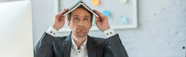 Disappointed businessman holding notebook on head with blurred mesh organizer on background, banner — Stock Photo