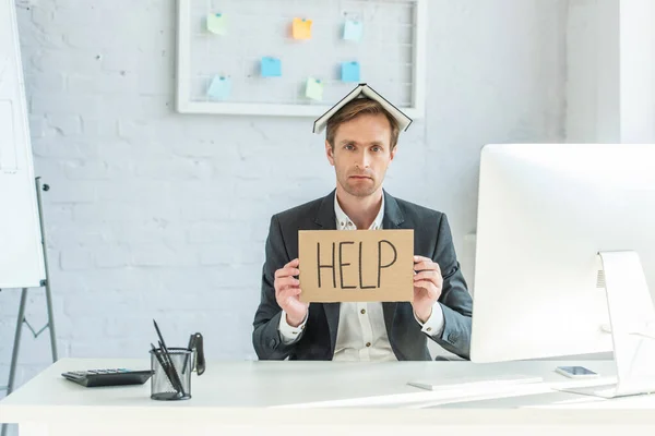 Sad businessman with notebook on head, holding cardboard with help lettering, while sitting at workplace — Stock Photo