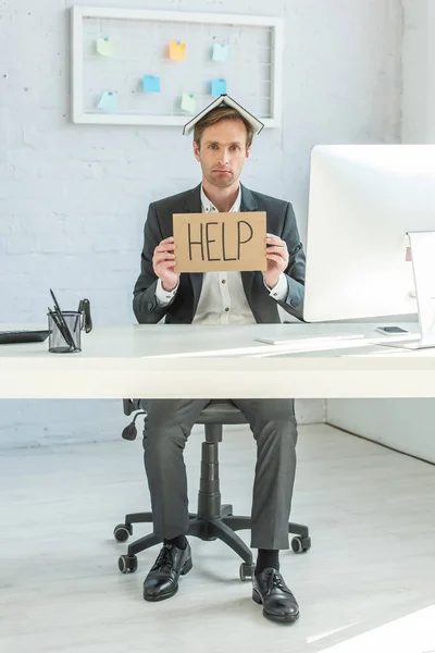 Tutta la lunghezza del fallimento deluso con notebook sulla testa, in possesso di cartone con scritte di aiuto, mentre seduto sul posto di lavoro — Foto stock