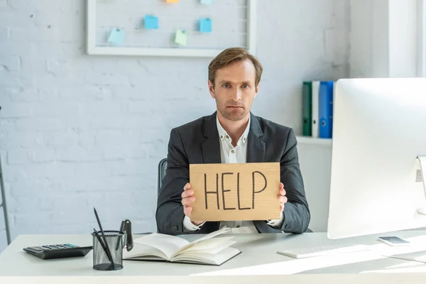 Sad businessman looking at camera and holding cardboard with help lettering, while sitting at workplace — Stock Photo