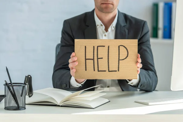 Cropped view of businessman holding cardboard with help lettering, while sitting at table on blurred background — Stock Photo