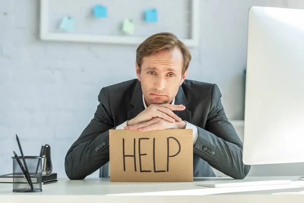 Disappointed businessman leaning on cardboard with help lettering, while sitting at workplace on blurred background — Stock Photo