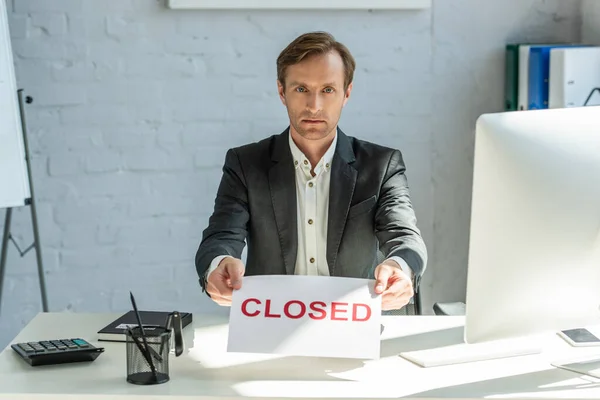 Serious businessman holding sign with closed lettering, while looking at camera at workplace — Stock Photo