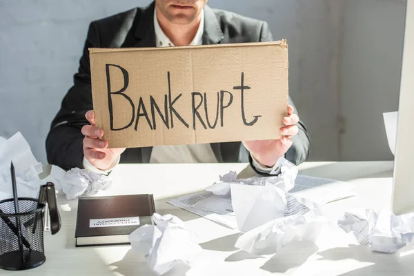 Cropped view of businessman holding cardboard with bankrupt lettering, while sitting at desk with crumpled papers — Stock Photo