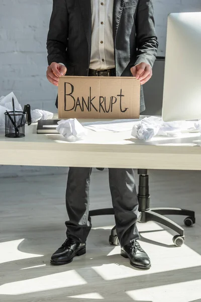 Cropped view of businessman holding cardboard with bankrupt lettering, while standing at workplace — Stock Photo