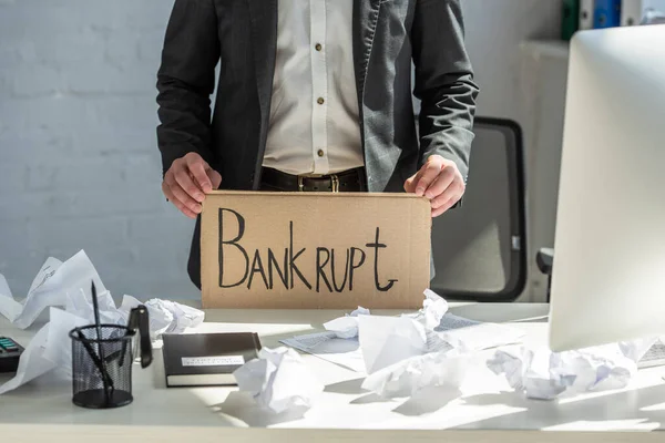 Cropped view of businessman holding cardboard with bankrupt lettering, while standing near table on blurred background — Stock Photo