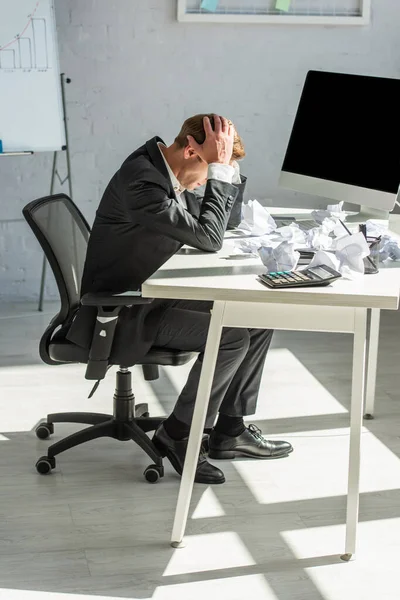 Full length of depressed businessman with hands on head, sitting at workplace with crumbled papers — Stock Photo