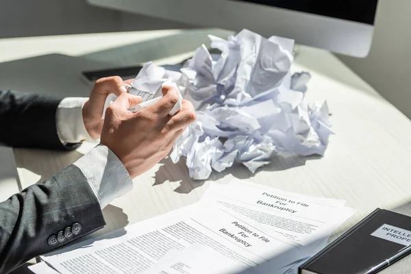 Cropped view of businessman holding crumbled paper near petitions for bankruptcy at workplace on blurred background — Stock Photo