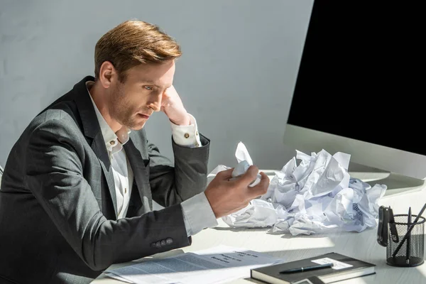 Disappointed businessman holding and looking at crumbled paper, while sitting at workplace with computer monitor — Stock Photo