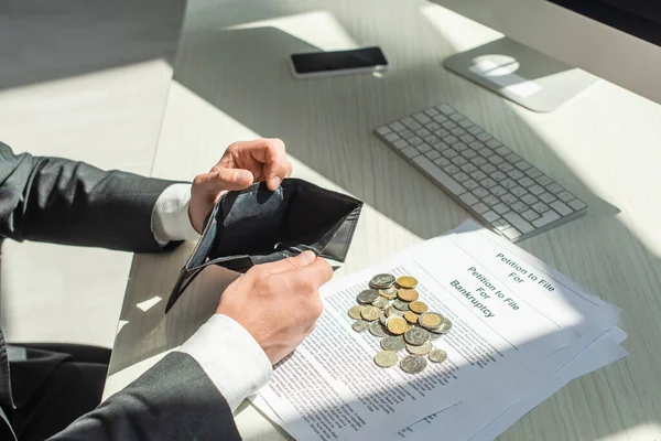 Обрізаний погляд бізнесмена, який тримає порожній гаманець біля монет і клопотання про банкрутство на робочому місці — стокове фото
