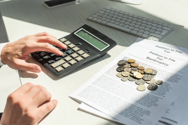 Vista recortada del hombre de negocios contando con calculadora cerca de monedas en peticiones de bancarrota en la mesa - foto de stock