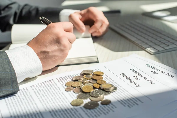 Vista recortada del hombre de negocios escribiendo en cuaderno cerca de monedas y peticiones de bancarrota con el lugar de trabajo borroso en el fondo - foto de stock