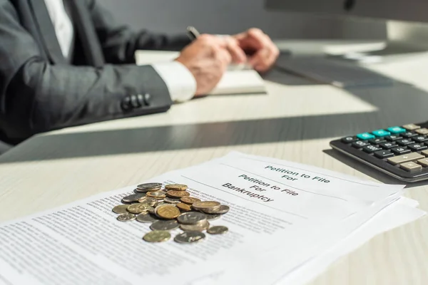 Vista recortada del hombre de negocios escribiendo en cuaderno cerca de monedas y peticiones de bancarrota sobre la mesa, sobre fondo borroso - foto de stock