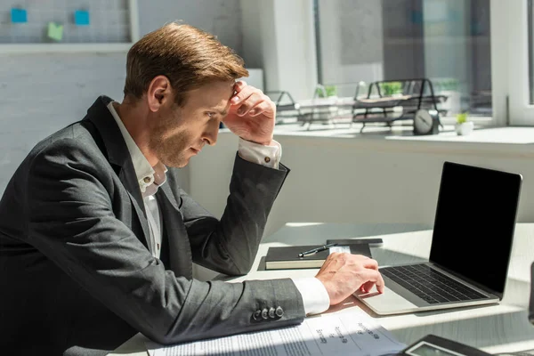 Hombre de negocios deprimido con la mano en la frente, utilizando el ordenador portátil, mientras está sentado en el lugar de trabajo con la oficina borrosa en el fondo - foto de stock