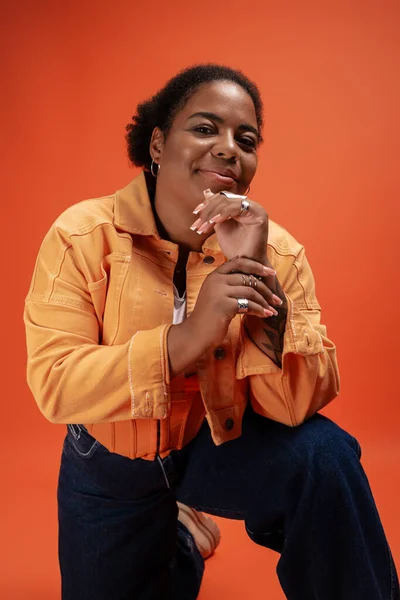 A trendy plus size woman poses confidently in a warm autumn outfit against a bold orange backdrop. — Stock Photo