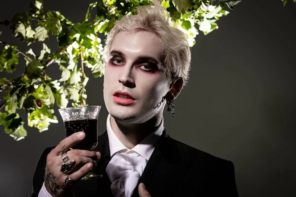Schöner junger Mann in einem aufwändigen Halloween-Kostüm hält ein dunkles Getränk in der Hand, das Charme versprüht. — Stockfoto