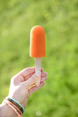 Hand holding orange popsicle on blurred green background clipart