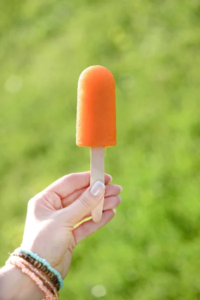 Hand Hält Orangefarbenes Eis Stiel Auf Verschwommenem Grünem Hintergrund — Stockfoto