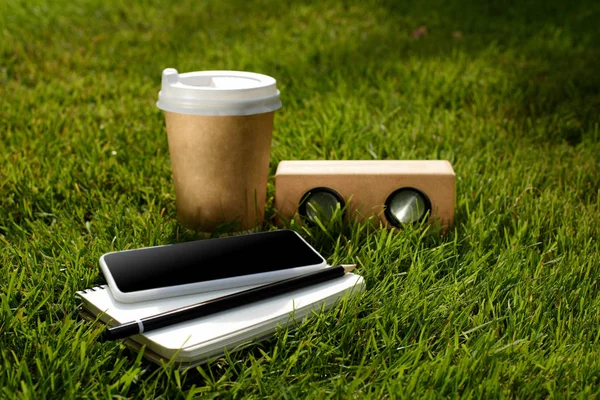 close up view of coffee to go, smartphone, notebook and audio speaker on green grass