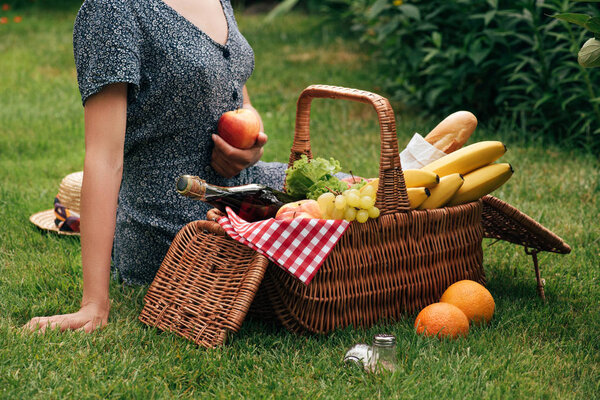 cropped image of woman sitting on green grass at picnic and holding ripe apple
