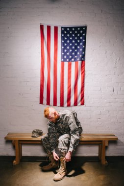 Amerikalı bir asker askeri üniformalı beyaz tuğla duvarın arkasında, 4 Temmuz tatil kavramı üzerinde bayrak ile ayakkabı bağcığı bağlama