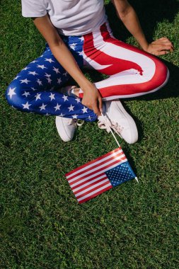 partial view of woman with flagpole in leggins with american flag pattern resting on green lawn, americas independence day holiday concept clipart