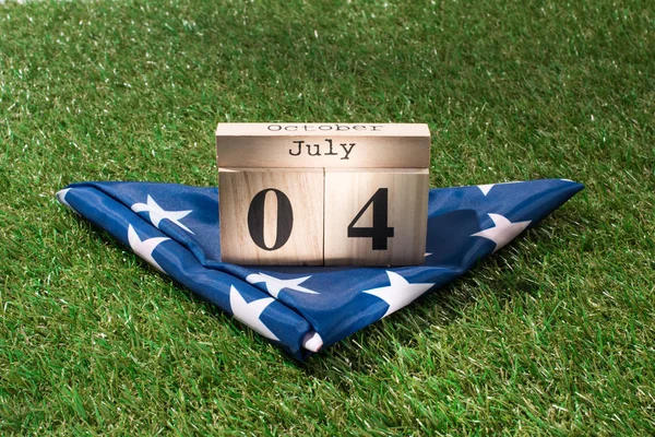 close up view of folded american flag and wooden calendar with 4th july date on green lawn, americas independence day concept