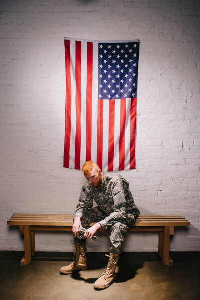 red hair american soldier sitting on wooden bench with flag on white brick wall behind, americas independence day concept